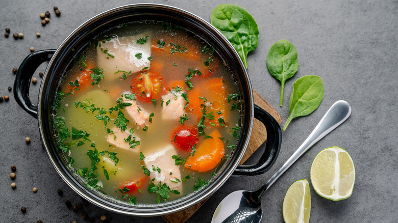 A pot of soup on a table top filled with chunks of meat, tomato, carrots, and green herbs