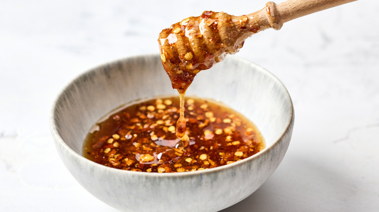 A honey spreader is dripping over a bowl of honey with chili flakes.