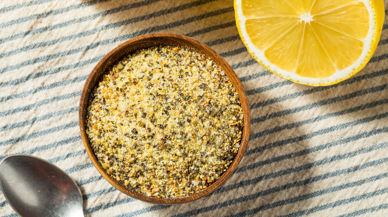 Close up of a bowl with lemon pepper seasoning next to a cut lemon and a spoon