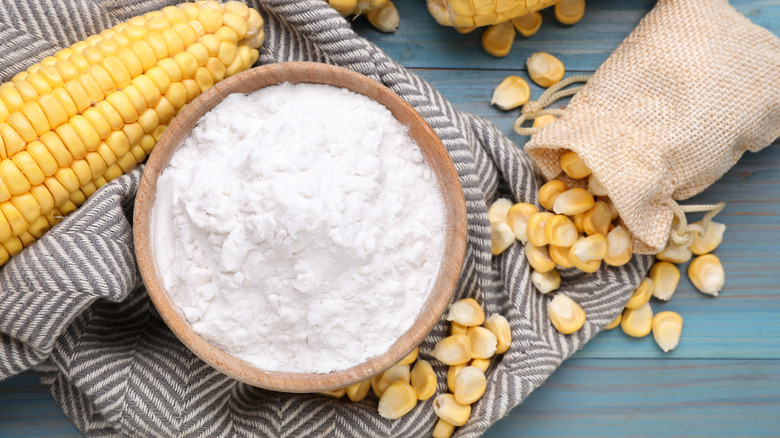 A bowl of cornstarch sits on a kitchen towel next to an ear of corn and a bag of spilled sweetcorn
