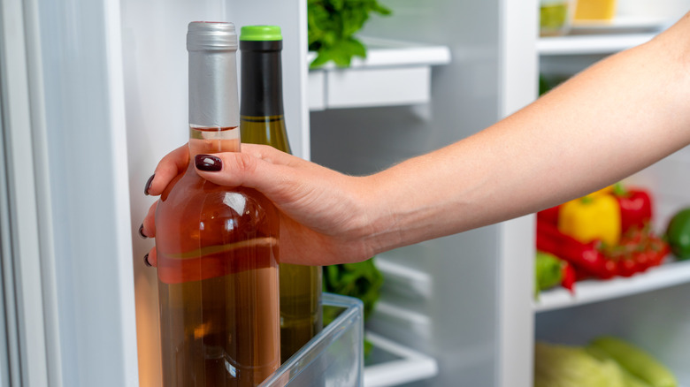 A hand reaches into the door shelf of an open fridge filled with produce and grasps a bottle of wine