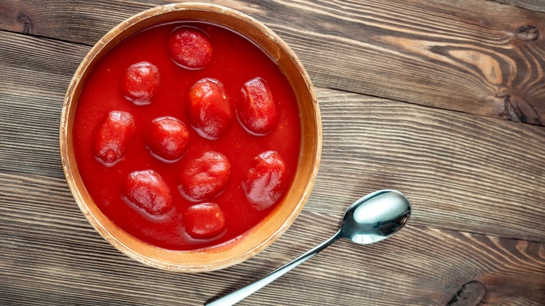 A bowl full of canned, whole tomatoes sitting on a wooden table next to a spoon