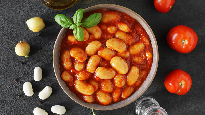 Round bowl of Italian style white beans in tomato sauce garnished with basil and surrounded by fresh tomatoes, garlic cloves, and white beans
