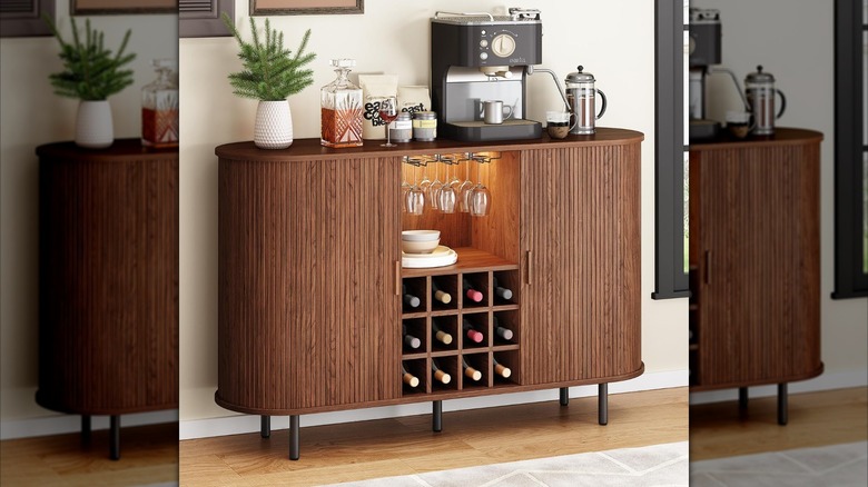 A walnut bar cabinet by the Onbrill brand against a white wall with various wine bottles and a coffee maker