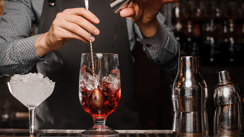 bartender making a drink with a stirrer in the glass with ice, another glass of ice, and a cocktail shaker on the bar.