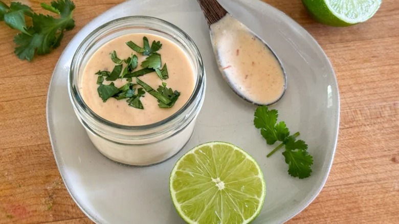 Chipotle crema in a jar resting on a plate, next to a spoon with crema, half a lime, and some spigs of cilantro