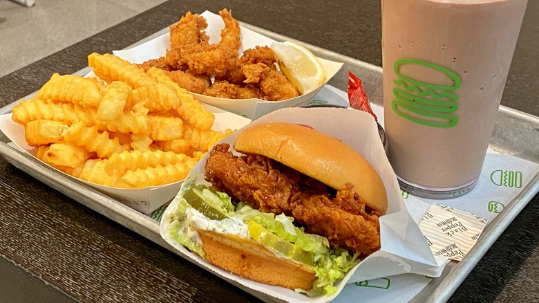 Food from Shake Shack on silver tray on top of dark table, including chicken sandwich, french fries, and chocolate milkshake.
