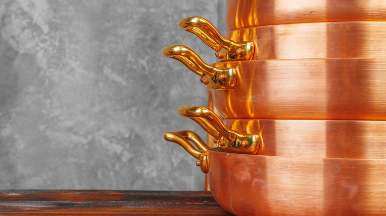 a nested stack of copper pots with brass handles, on a wooden shelf with a grey wall to the rear