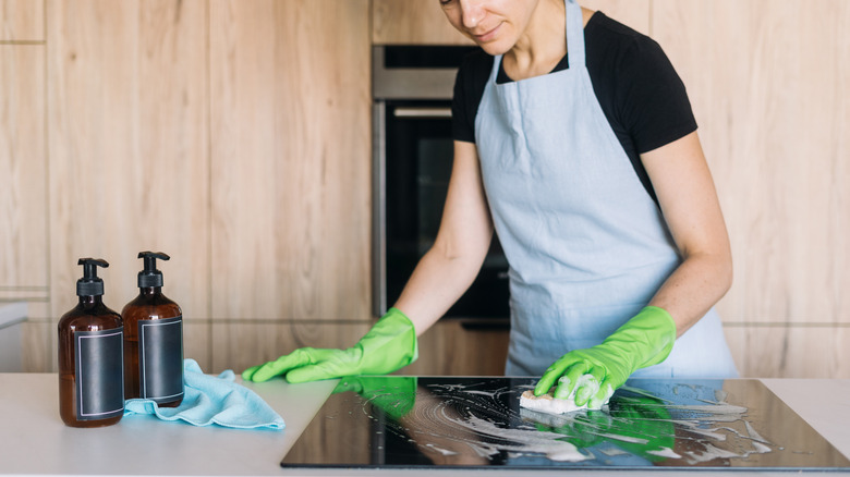 young woman in black T-shirt, blue apron and green gloves scrubbing an induction cooktop in a sleek, Scandinavian-looking kitchen