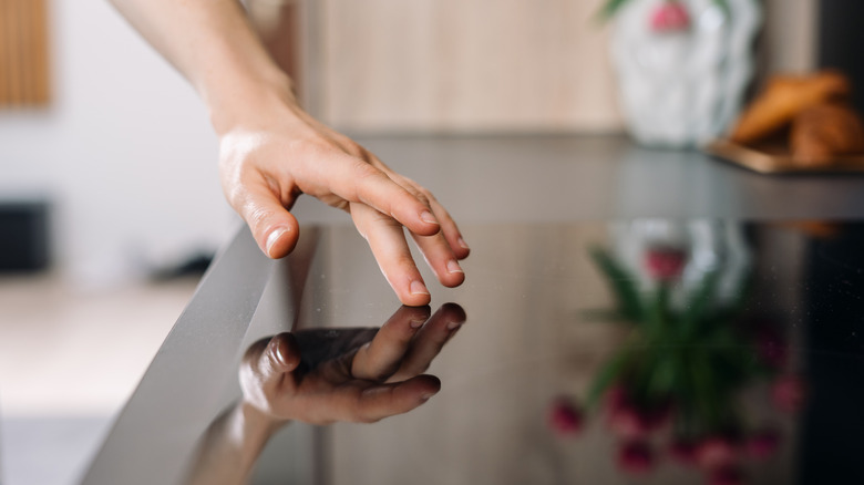 a hand lightly touching a shiny stovetop with one finger, in a sleek and modern-looking kitchen with a vase of flowers visible in the background and also as a reflection in the stovetop