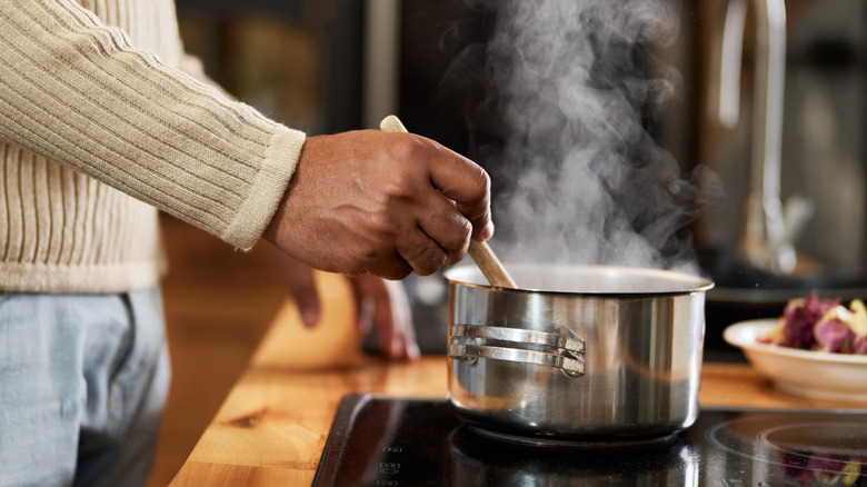A man's hand holding a wooden spoon and stirring a pot with steam coming out of it