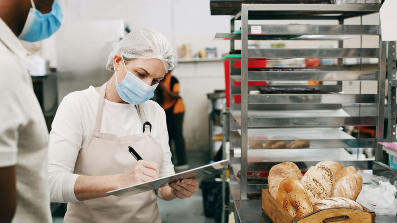 a health inspector in a mask and hairnet taking notes on a clipboard in a bakery