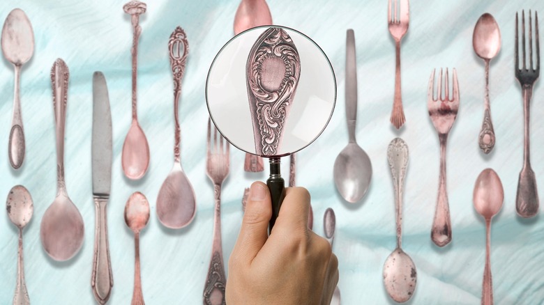 A hand holding a magnifying glass over silver cutlery