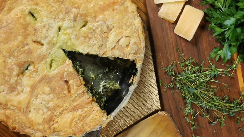 overhead view of spinach pie with slice missing beside cutting board of herbs and cheese