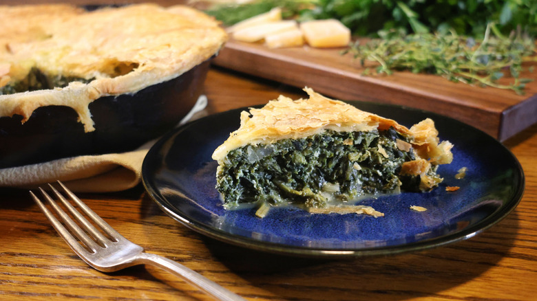 plated slice of spinach pie beside cast iron skillet and cutting board with cheese and herbs