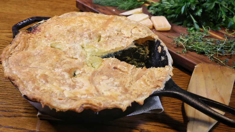 cast iron skillet spinach pie beside wooden spatula and cutting board with cheese and herbs