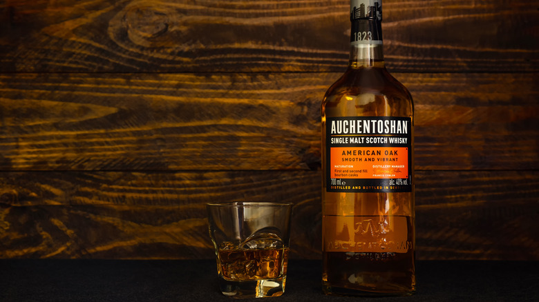 A bottle of Auchentoshan American Oak in front of a dark wooden background next to a filled glass