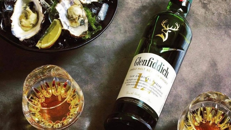 A bottle of Glenfiddich 12-year laid on its back next to filled glasses and a plate of seafood