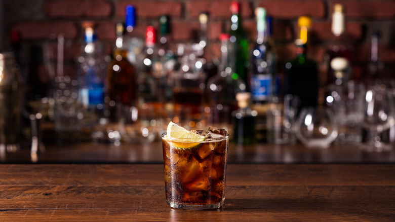 A cola cocktail with a slice of lime placed on a bar with blurred bottles in the background