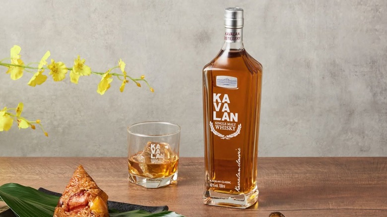 A bottle and filled glass of Kavalan Classic posed on a wooden surface next to food and a plant