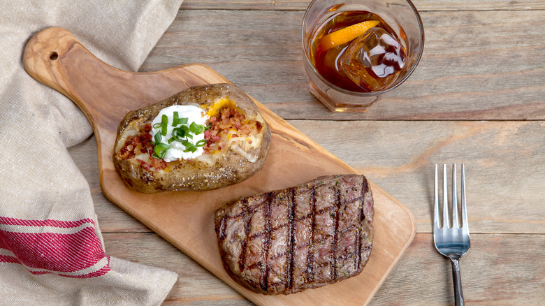 Steak and baked potato on cutting board with whiskey cocktail