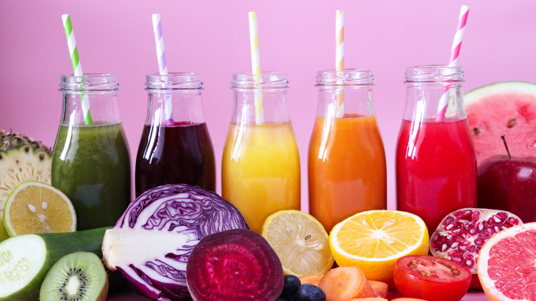 Five glass bottles of juice with straws surrounded by fresh fruits and vegetables, including green, purple, yellow, orange, and red varieties