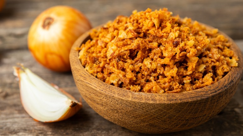 A wooden bowl of crispy fried onions next to a quartered raw onion and a whole unpeeled onion.