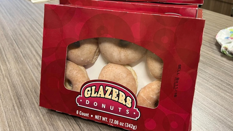 A red box of Glazers donuts upright on a wood grain table with more boxes behind it