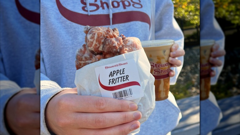 A hand holds an apple fritter wrapped in wax paper and a Stewart's iced coffee