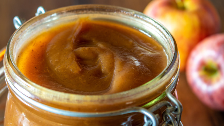 A jar of homemade apple butter with red apples seen in the background