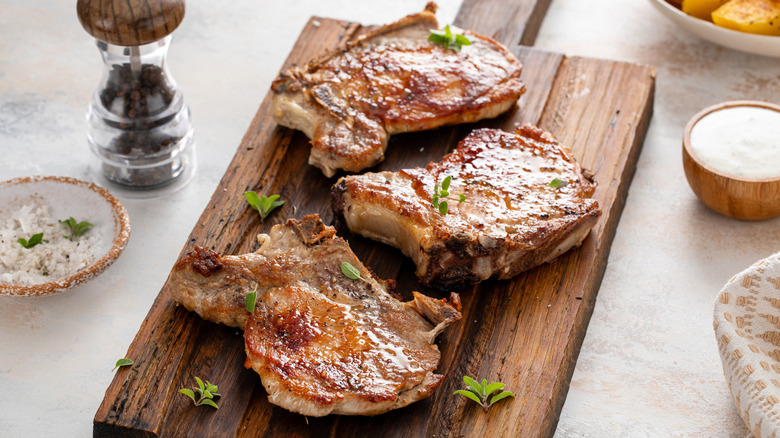 Cooked pork chops on a wooden cutting board with a pepper shaker and bowl of salt nearby