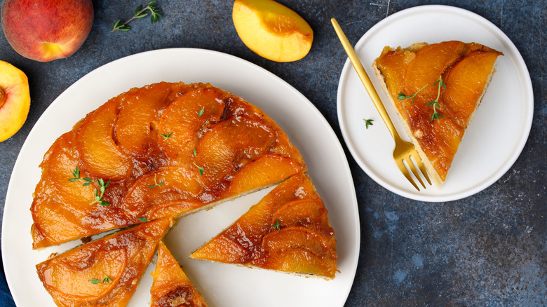 A peach upside-down cake on a white plate on a blue tablecloth, next to a slice of the cake on a smaller plate with a fork, surrounded by sliced and whole peaches