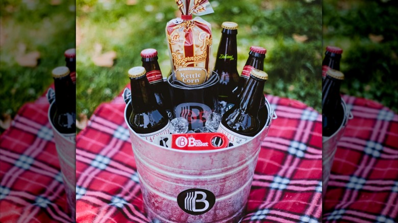 Bottled beer packaged in a metal bucket sitting on a plaid blanket on the ground
