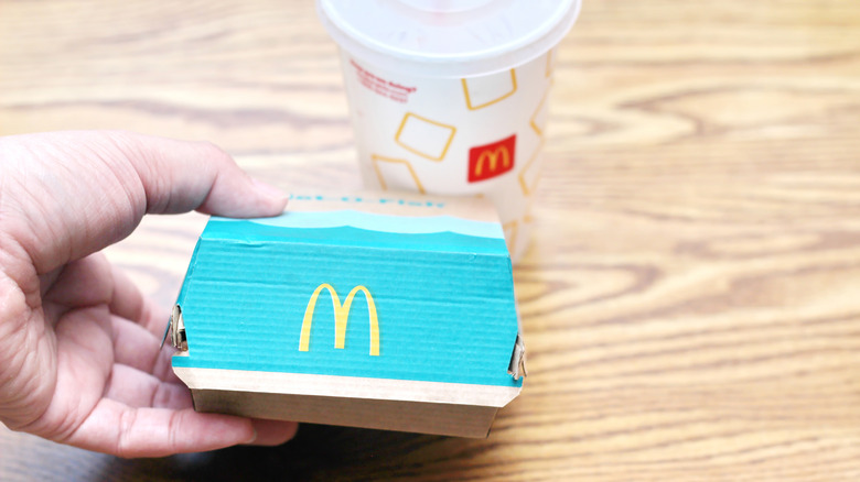 A hand holding and lifting a Filet-O-Fish, with a McDonalds cup in the background