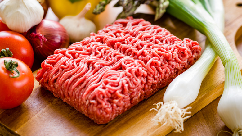Block of raw ground beef on wooden cutting board next to vegetables including tomatoes, leeks, and onions