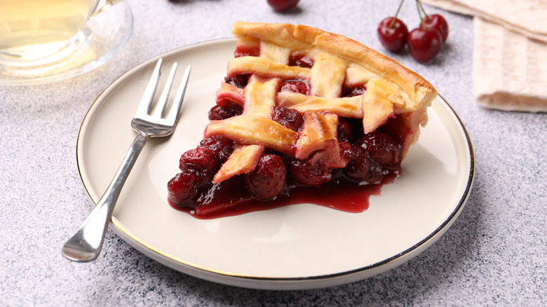 Slice of cherry pie on white plate with silver fork next to fresh cherries