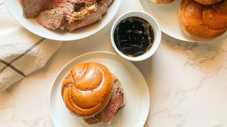 Sliced beef sandwich on a plate next to a bowl of beef drippings