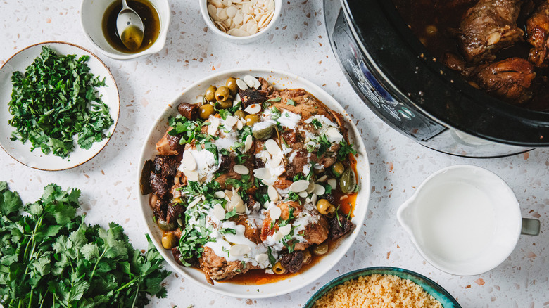 Large plate with bone-in chicken thighs with olives, figs, yogurt and cilantro, surrounded by more toppings and a slow cooker with chicken parts