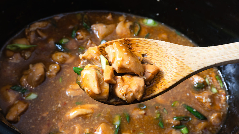 Wooden spoon lifting chicken pieces in orange sauce above slow cooker
