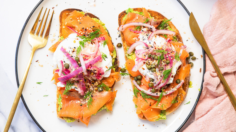 Two slices of smoked salmon breakfast toast on white plate with cutlery.