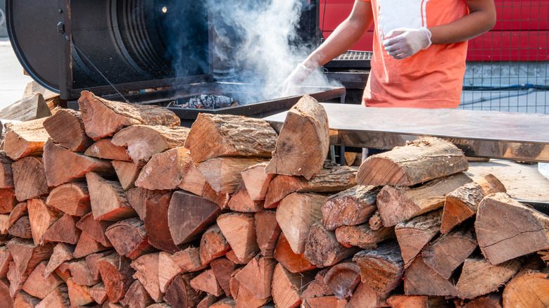 variety of woods next to smoker billowing smoke and man berbecueing