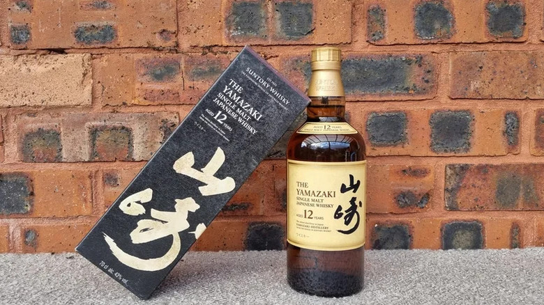 A bottle of Yamazaki 12-year in front of a red brick background with its box leaning against it