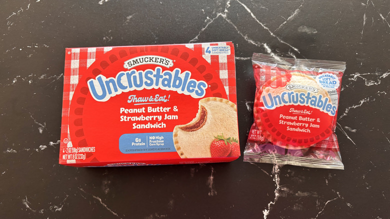 Smucker's Uncrustables box next to Uncrustable sandwich in wrapper on a marbled black counter