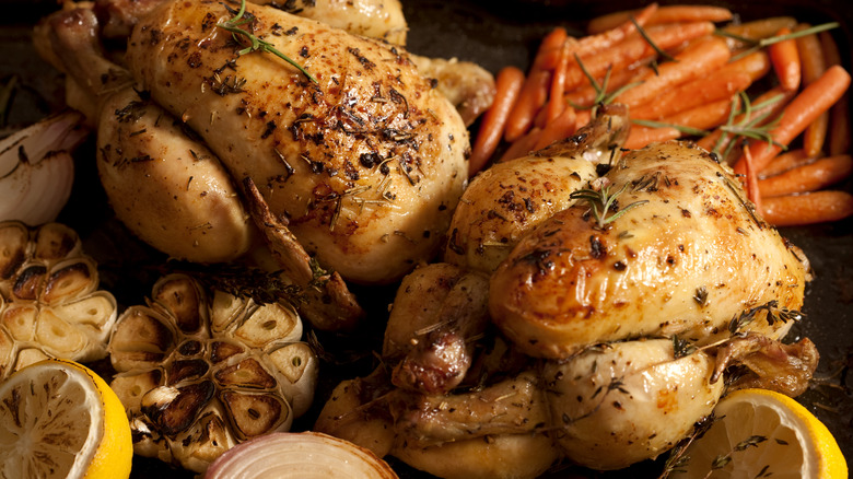 Two whole roasted chickens surrounded by carrots, lemons, and garlic heads.