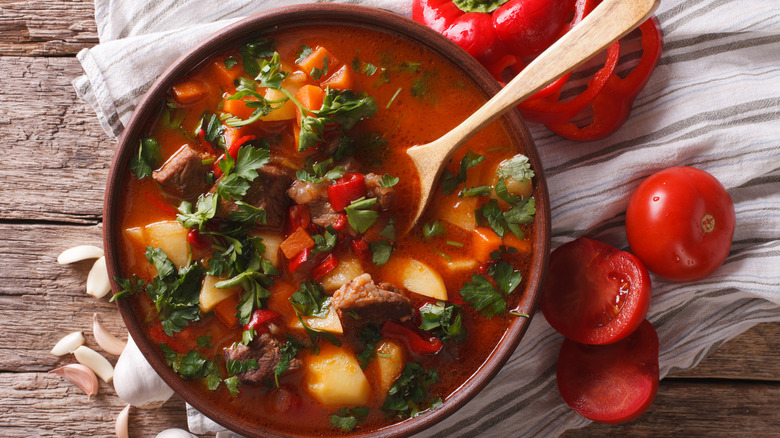A pot of red soup full of herbs and vegetables with a wooden spoon in it, sitting on a cloth next to some red peppers and tomatoes