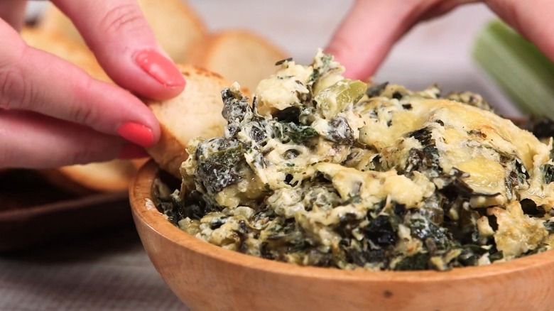 Hand dunking crostini into bowl of cheesy collard greens dip