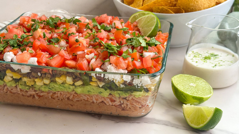 7-layer dip in glass baking dish, next to lime wedges and jug of creamy dressing