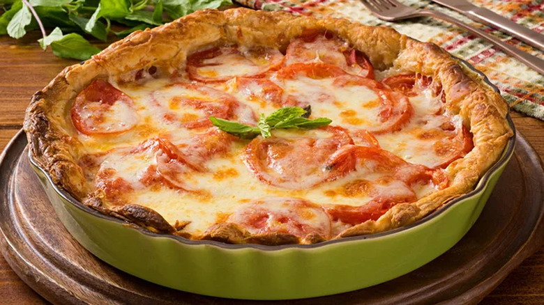A warm, cheesy tomato pie in a green pie dish topped with a sprig of basil