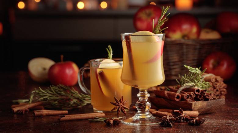 Two apple cider cocktails on a wooden table surrounded be rosemary sprigs, spices, and apples