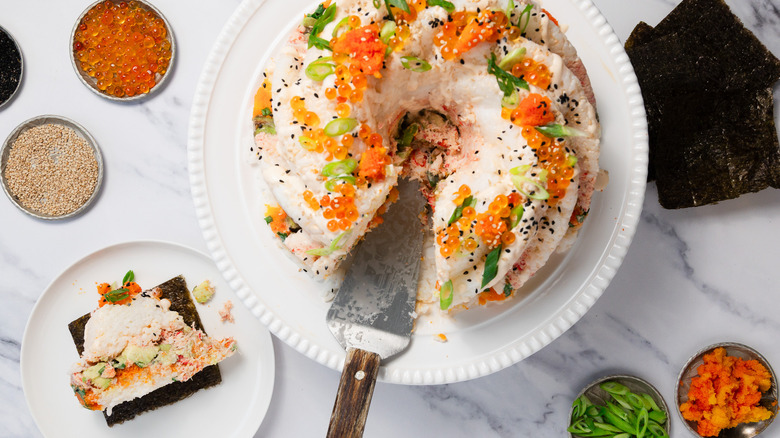 sushi bundt cake on cake stand with nori and toppings, besides a slice taken out
